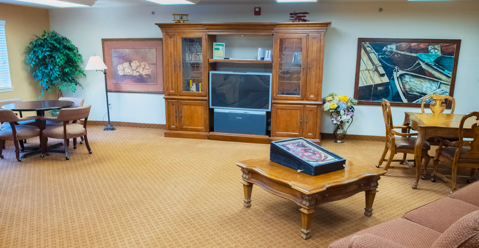 A cozy living room area with a wooden entertainment center holding a television, a wooden coffee table with a game on top, a round table with four chairs, a sofa, two large framed paintings on the walls, a floor lamp, and a potted plant in the corner.
