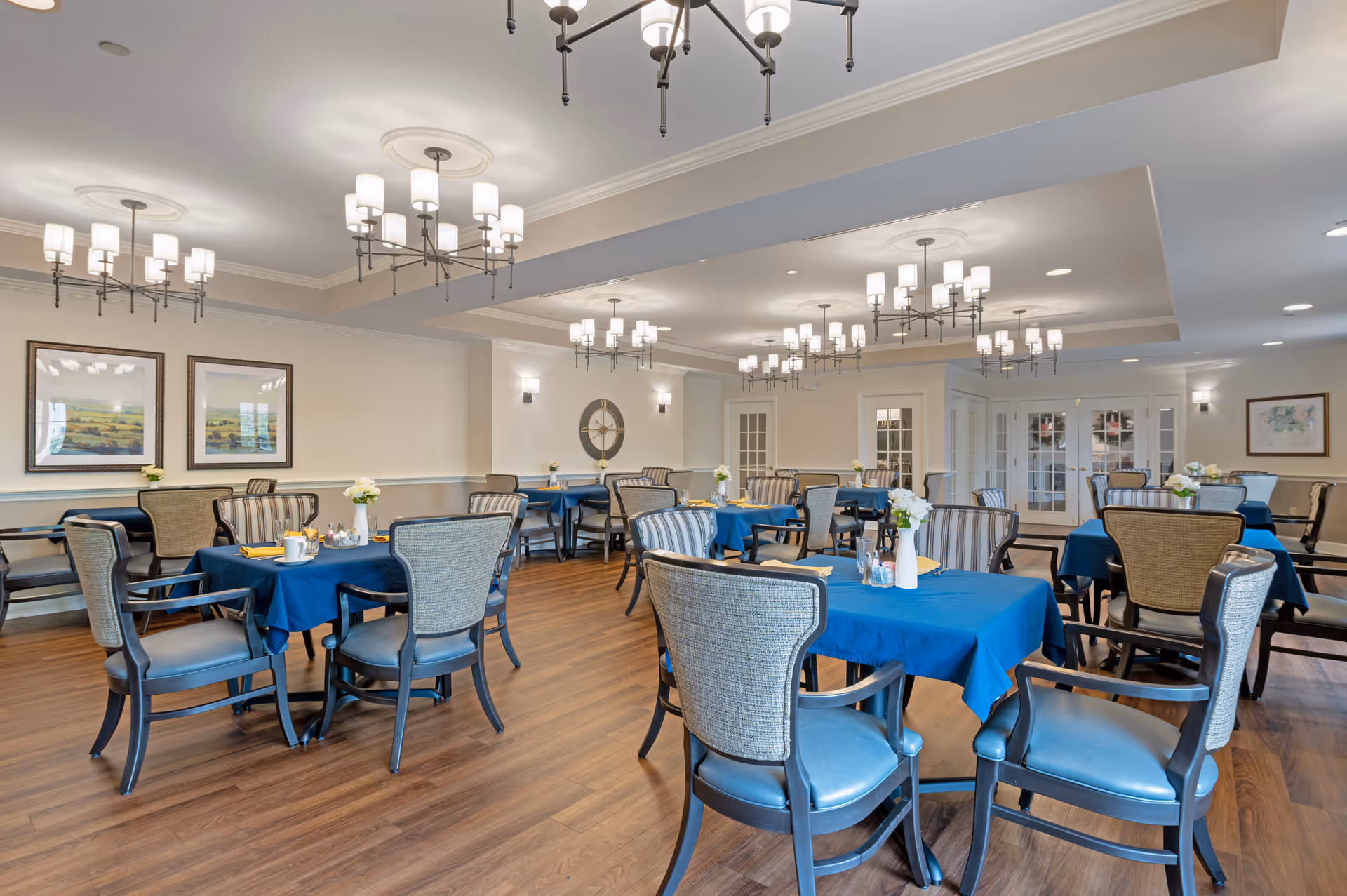 A spacious dining room with multiple tables covered in blue tablecloths, each set with chairs and small flower vases. The room features wooden flooring, elegant chandeliers, framed artwork on the walls, and large windows allowing natural light.