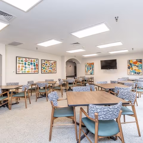 A bright dining room with multiple wooden tables and chairs featuring patterned cushions. The walls are decorated with colorful abstract paintings, and a flat-screen TV is mounted on one wall. The ceiling has several recessed lights, and the room has a clean, modern look.