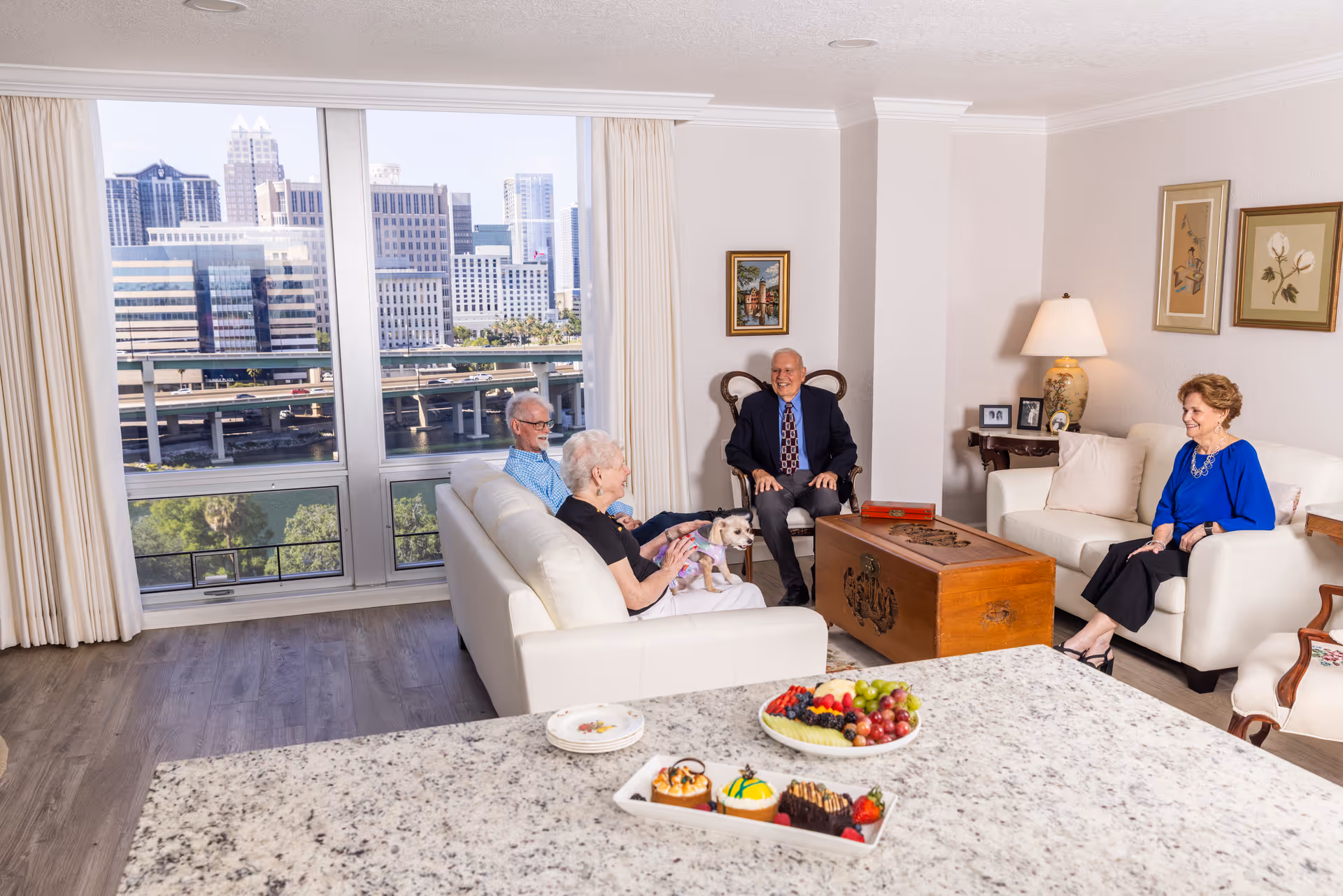 Four older adults and a small dog sitting and chatting in a bright living room with a city skyline visible through large windows and a kitchen counter with fruit in the foreground.