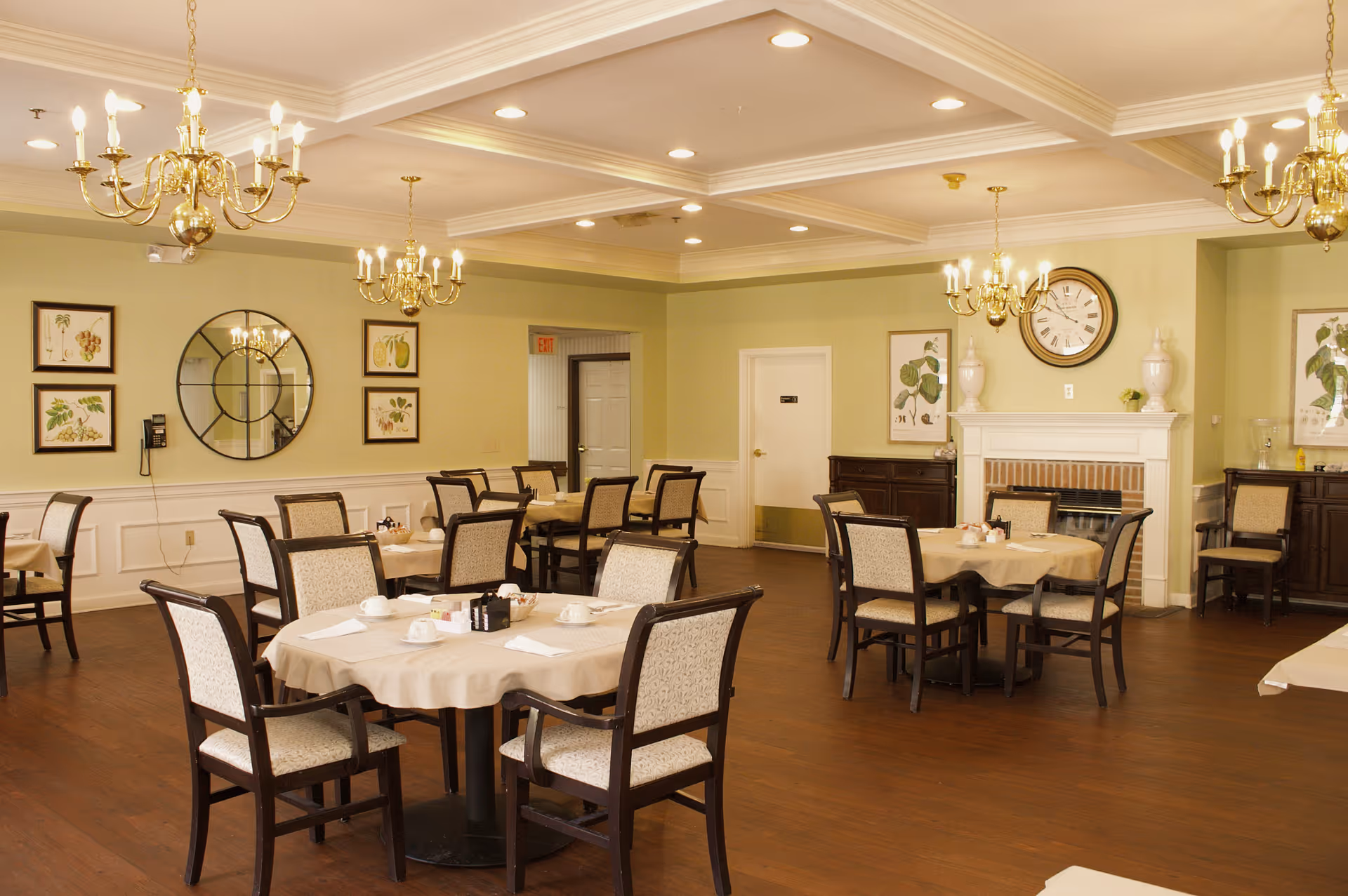 A dining room with several round tables covered with beige tablecloths, each surrounded by four upholstered chairs. The room features wooden flooring, light green walls with white wainscoting, framed botanical prints, a large round mirror, and a fireplace with a clock above it. Multiple brass chandeliers hang from the ceiling providing warm lighting.