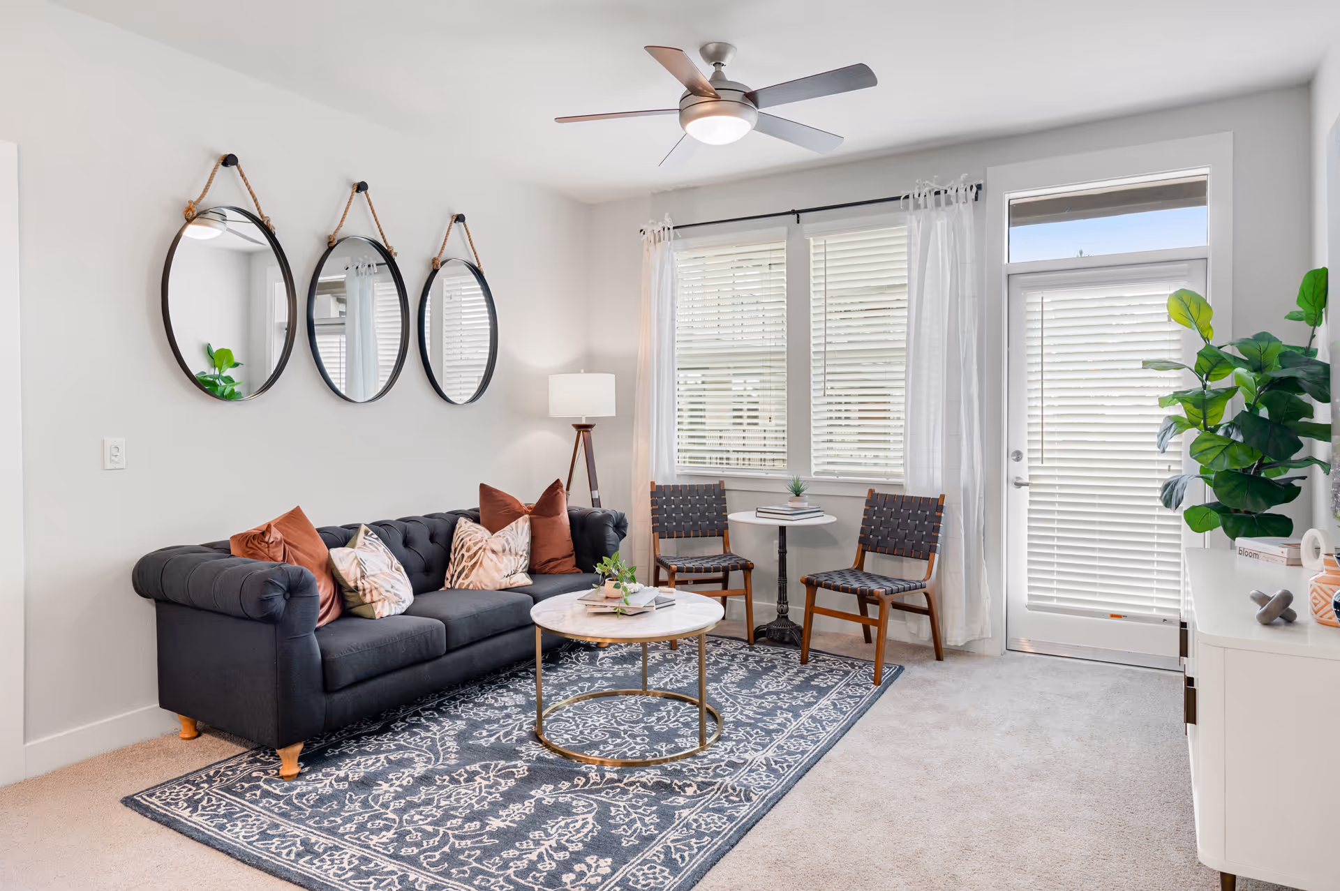 Bright modern living room with a dark tufted sofa and cushions, three round wall mirrors, a coffee table on a patterned rug, and chairs by a windowed door.