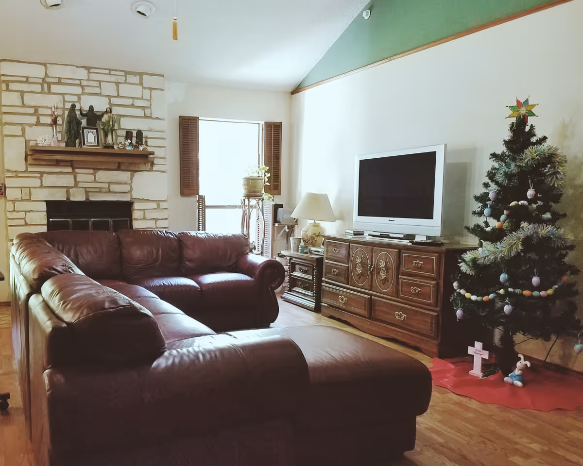 A cozy living room with a large brown leather sectional sofa facing a wooden cabinet with a flat-screen TV on top. To the right of the TV is a decorated Christmas tree with ornaments and a star on top. On the left side of the room is a stone fireplace with a wooden mantle displaying religious statues and decorative items. A window with wooden shutters lets in natural light behind the sofa.