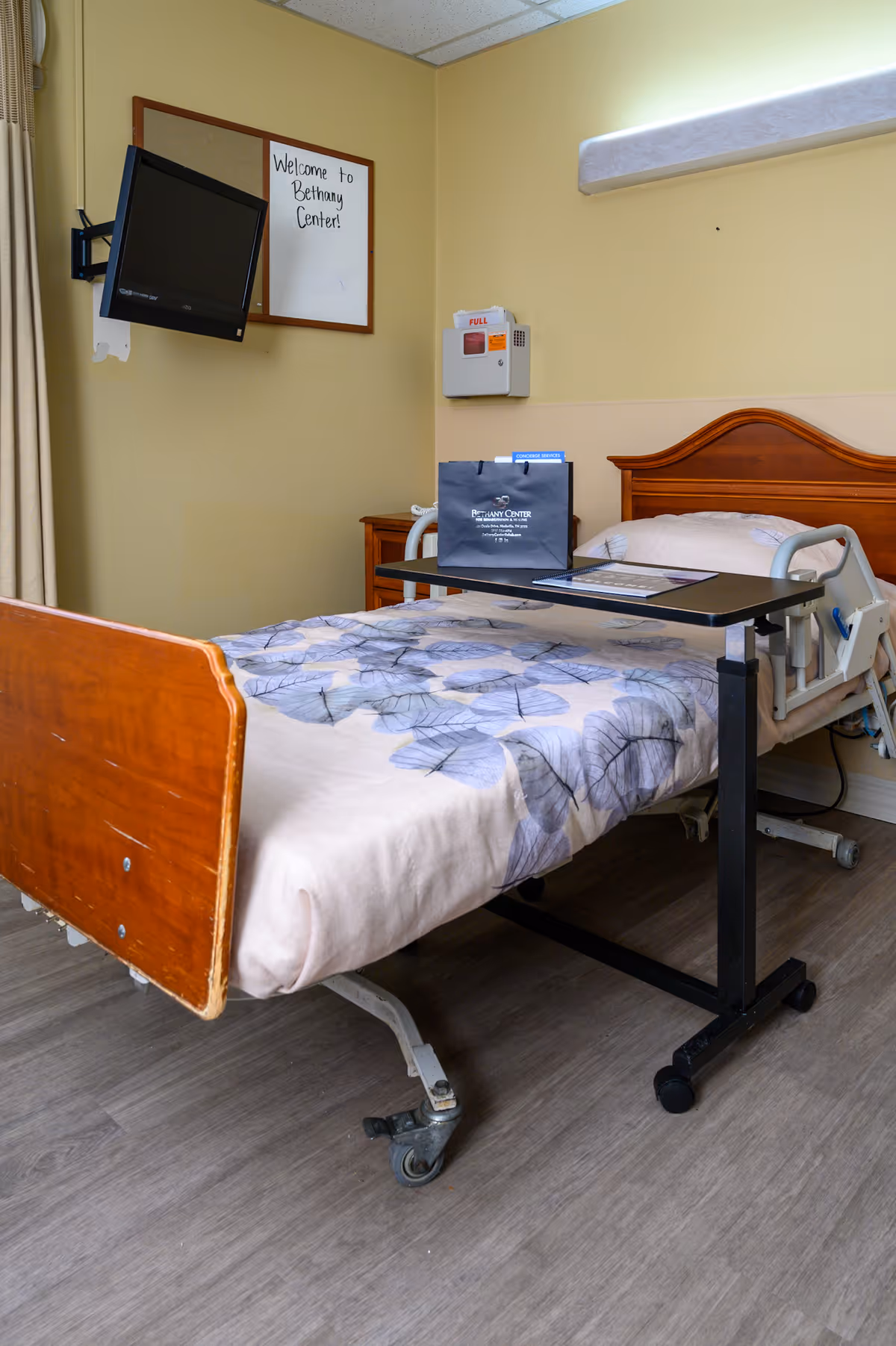 A hospital-style bed with a floral patterned blanket in a room with light yellow walls. A small overbed table holds a dark blue bag with 'Bethany Center for Rehabilitation and Healing' printed on it. A wall-mounted TV is angled towards the bed, and a whiteboard on the wall reads 'Welcome to Bethany Center!'.