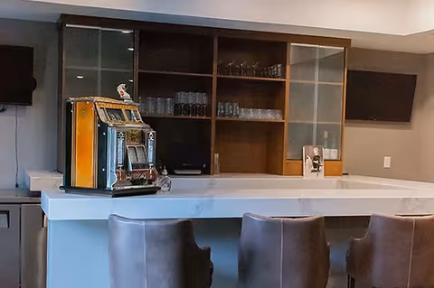 A modern bar area with a white countertop and three brown leather bar stools. Behind the bar is a wooden shelving unit with glassware and a vintage slot machine on the counter. Two flat-screen TVs are mounted on the walls on either side of the shelving unit.
