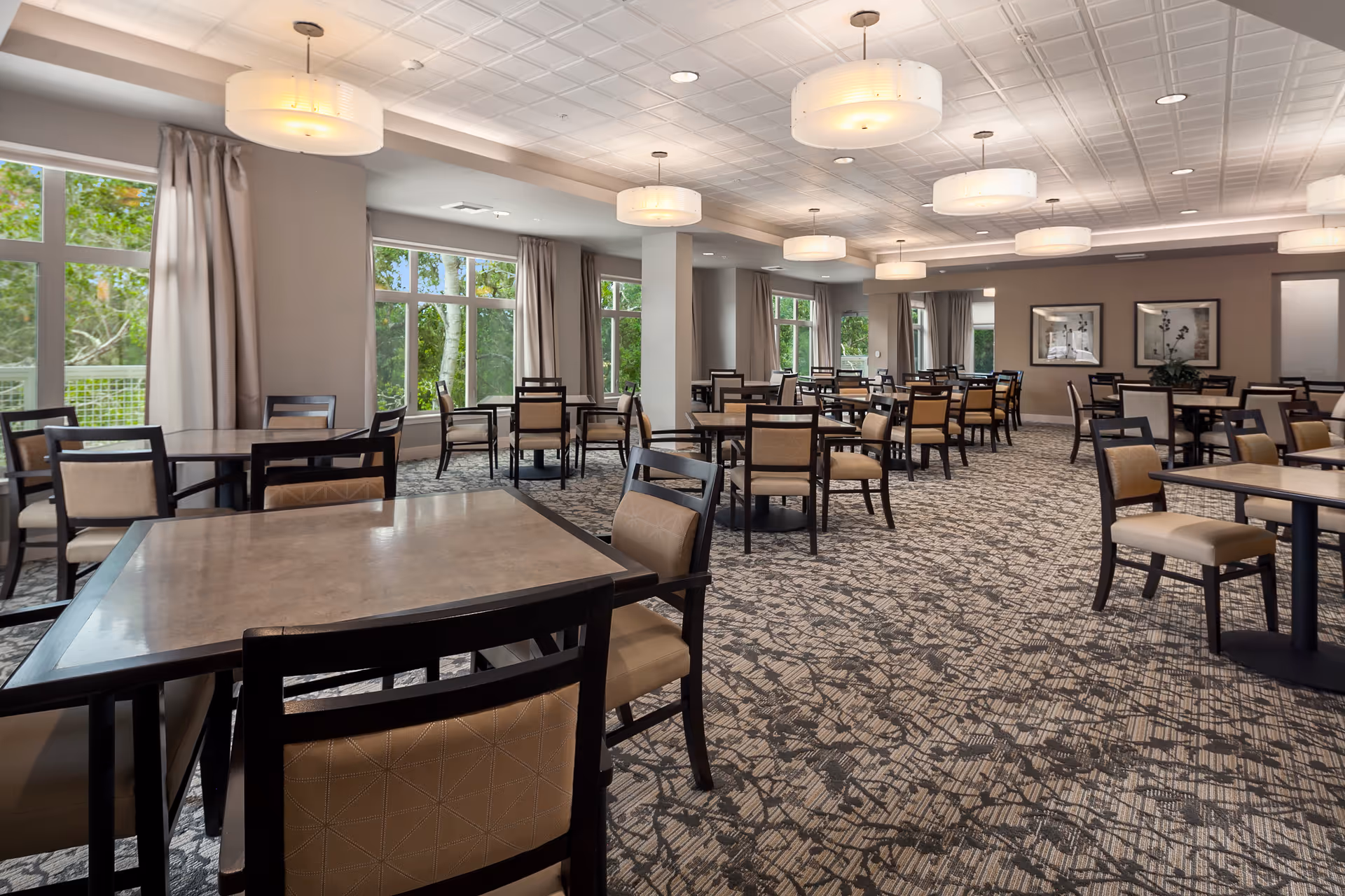 A spacious dining room with multiple square tables and cushioned chairs arranged neatly. Large windows with beige curtains allow natural light to fill the room, and modern ceiling lights hang from a white tiled ceiling. The carpet has a patterned design, and framed artwork is visible on the far wall.