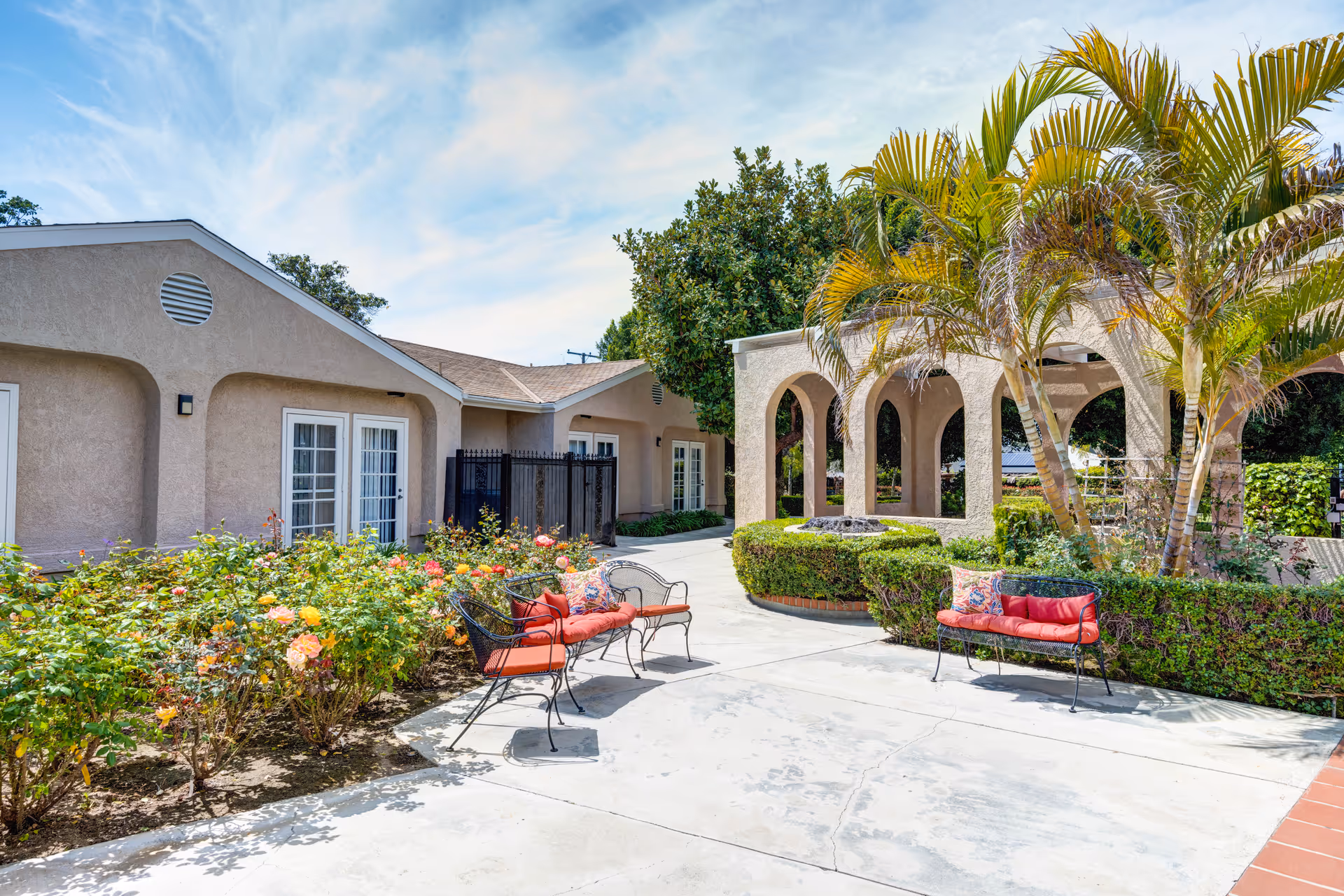 Sunny courtyard with red-cushioned benches, rose bushes, palm trees, and an arched colonnade beside a beige building.