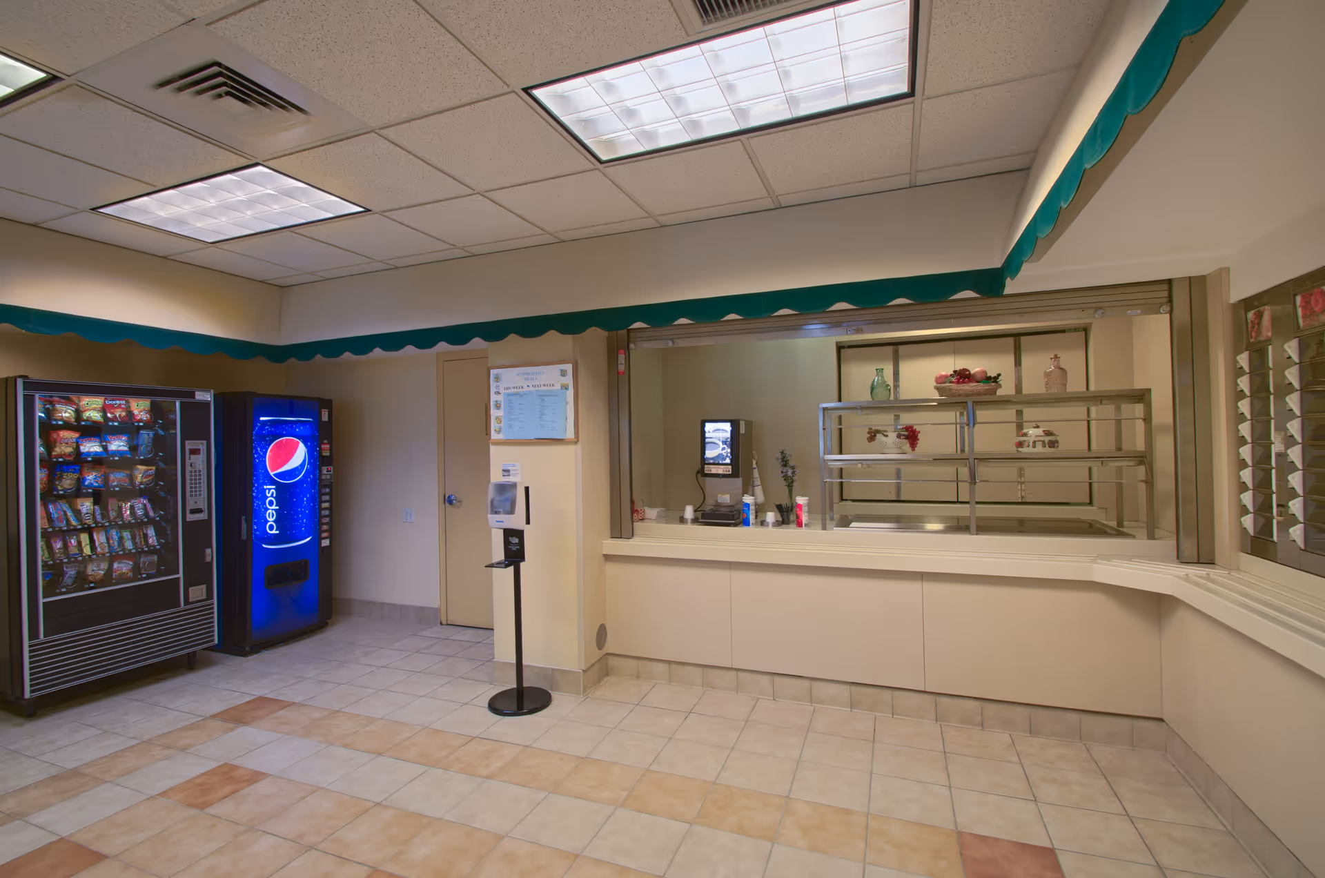A tiled room with snack and Pepsi vending machines and a food service counter with a pass-through window.