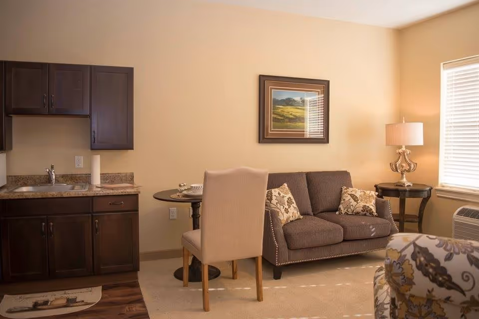 A cozy senior living room area with a small kitchenette on the left featuring dark wood cabinets and a sink. In the center, there is a round dining table with a beige upholstered chair. To the right, a dark gray loveseat with two floral cushions sits beneath a framed landscape painting. A side table with a decorative lamp is next to the loveseat, and a window with blinds allows natural light into the room. A patterned armchair is partially visible in the foreground.