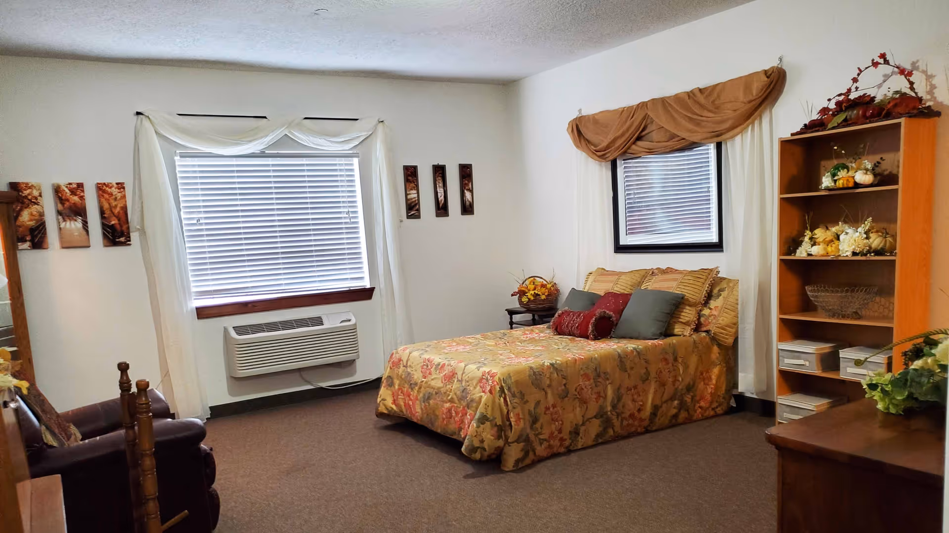 A cozy bedroom with a floral bedspread on a double bed, multiple pillows, a wooden bookshelf filled with decorative items, a wooden dresser with a plant, two windows with blinds and curtains, and a brown armchair in the corner.