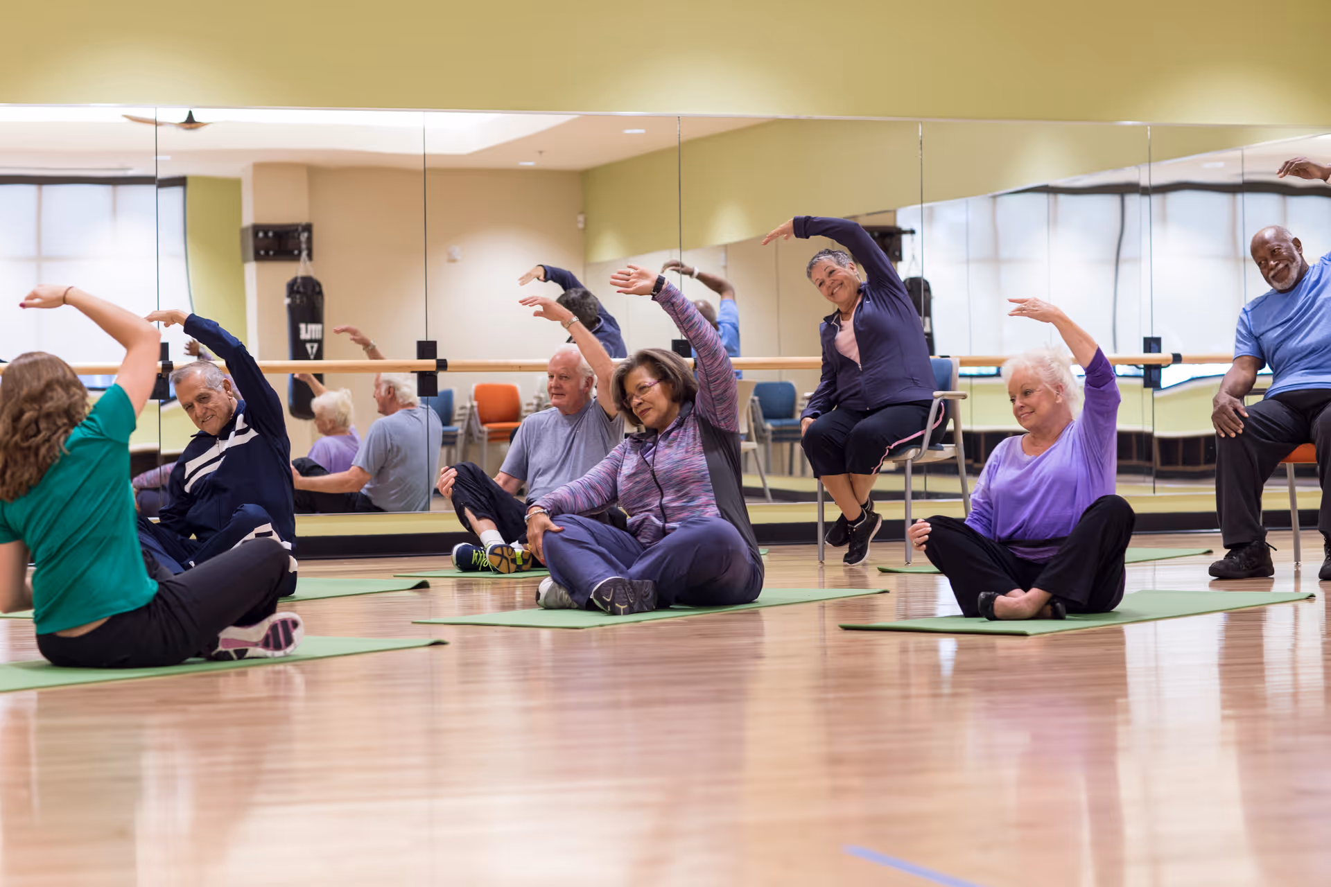 A group of elderly people participating in a seated stretching exercise class in a fitness room with wooden floors and large mirrors on the wall. Some participants are sitting on green exercise mats on the floor, while one person is seated on a chair. They are all raising one arm overhead and leaning to the side.