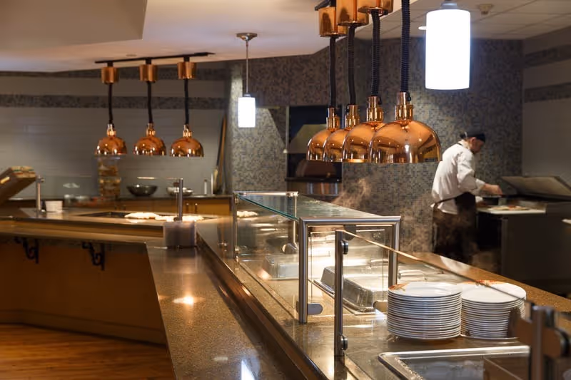 Cafeteria-style serving line with copper heat lamps, a glass sneeze guard, stacked plates, and a chef working in the kitchen.