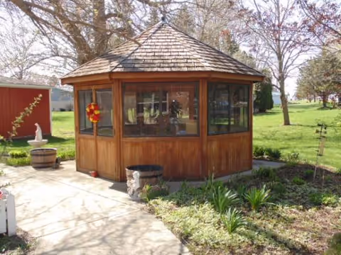 Wooden gazebo with glass windows sits in a well-kept garden near a paved path.