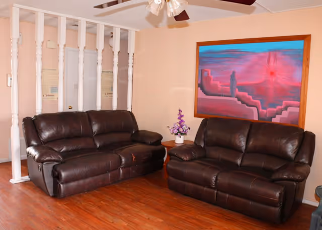 A cozy living room area with two dark brown leather sofas facing each other on a wooden floor. Between the sofas is a small round wooden table with a vase of purple flowers. The wall behind one sofa features a large colorful painting with abstract shapes in shades of pink, purple, and blue. White vertical wooden spindles partially separate this area from an adjacent space.