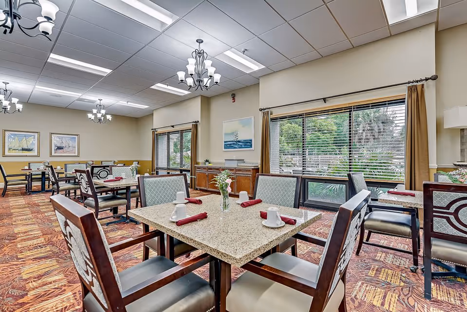 A dining room in a senior living facility with several tables and chairs arranged neatly. Each table has a granite top, set with white cups and red napkins. The room features large windows with brown curtains, allowing natural light to brighten the space. The walls are decorated with framed artwork, and the ceiling has multiple chandeliers and fluorescent lights. The carpet has a patterned design in warm tones.