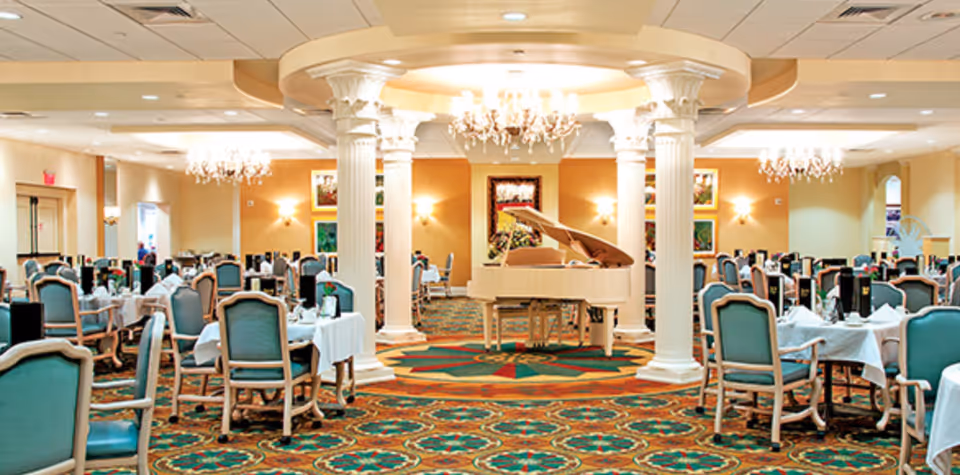 A spacious dining room with multiple tables covered in white tablecloths and set with napkins and tableware. The room features elegant chandeliers, decorative columns, and a white grand piano in the center. The carpet has a colorful geometric pattern, and the walls are adorned with framed artwork and warm lighting.