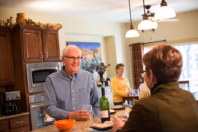 An elderly man and woman are socializing in a kitchen area. The man is standing behind a kitchen counter holding a wine glass and smiling, while the woman is seated on the other side of the counter. In the background, another elderly woman is standing near a dining table with wine glasses. The kitchen has wooden cabinets, a microwave, a coffee maker, and pendant lights hanging from the ceiling.