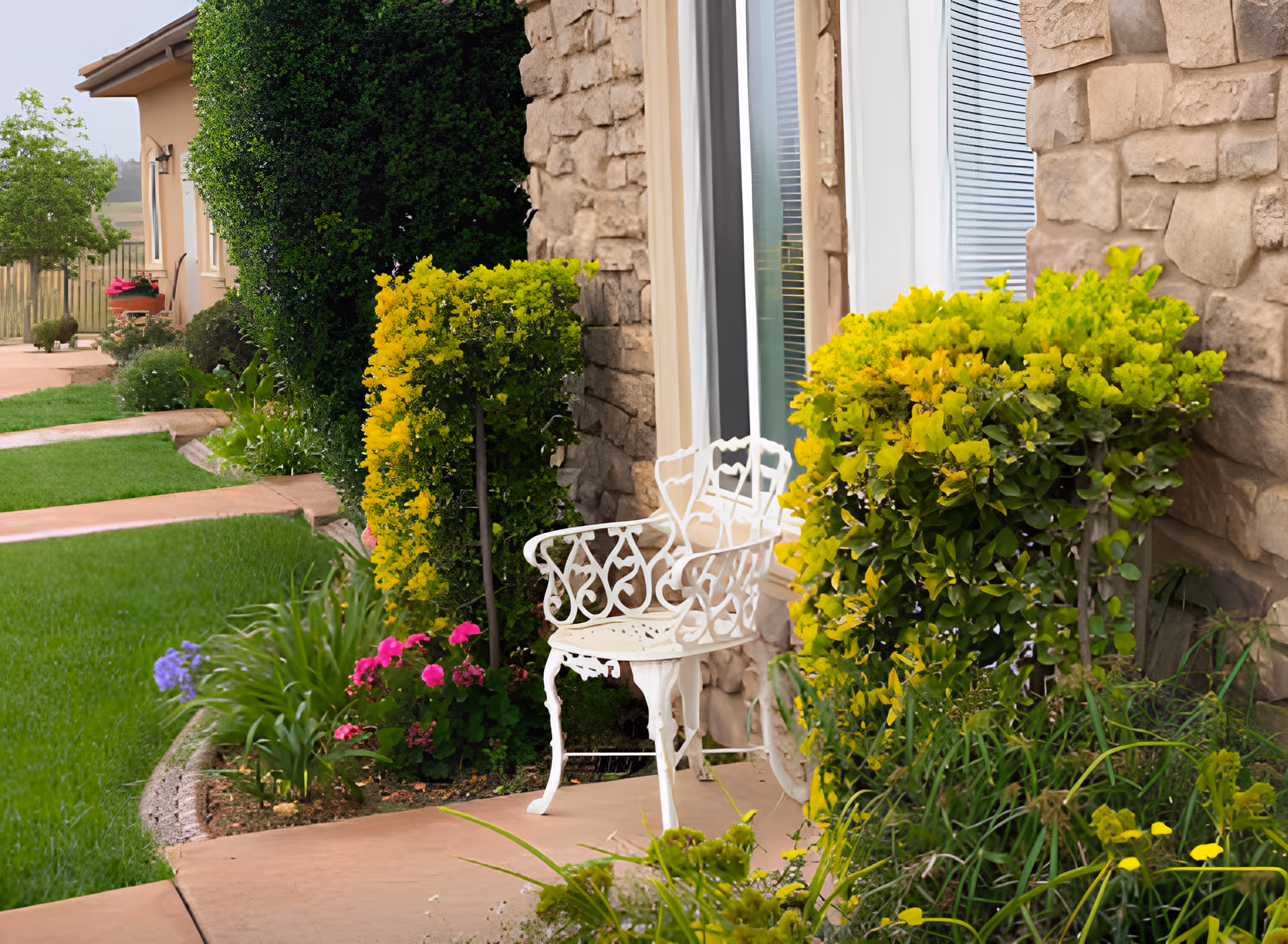 A white decorative metal chair sits on a stone patio next to a window with blinds. The patio is surrounded by lush green and yellow bushes, colorful flowers, and well-maintained grass. The exterior wall of the building is made of stone, and a pathway leads along the side of the building with more greenery and plants visible.