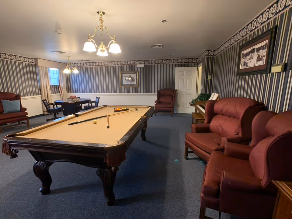 A senior living common room with a pool table in the center, red upholstered chairs, a small table and framed pictures against striped wallpaper.