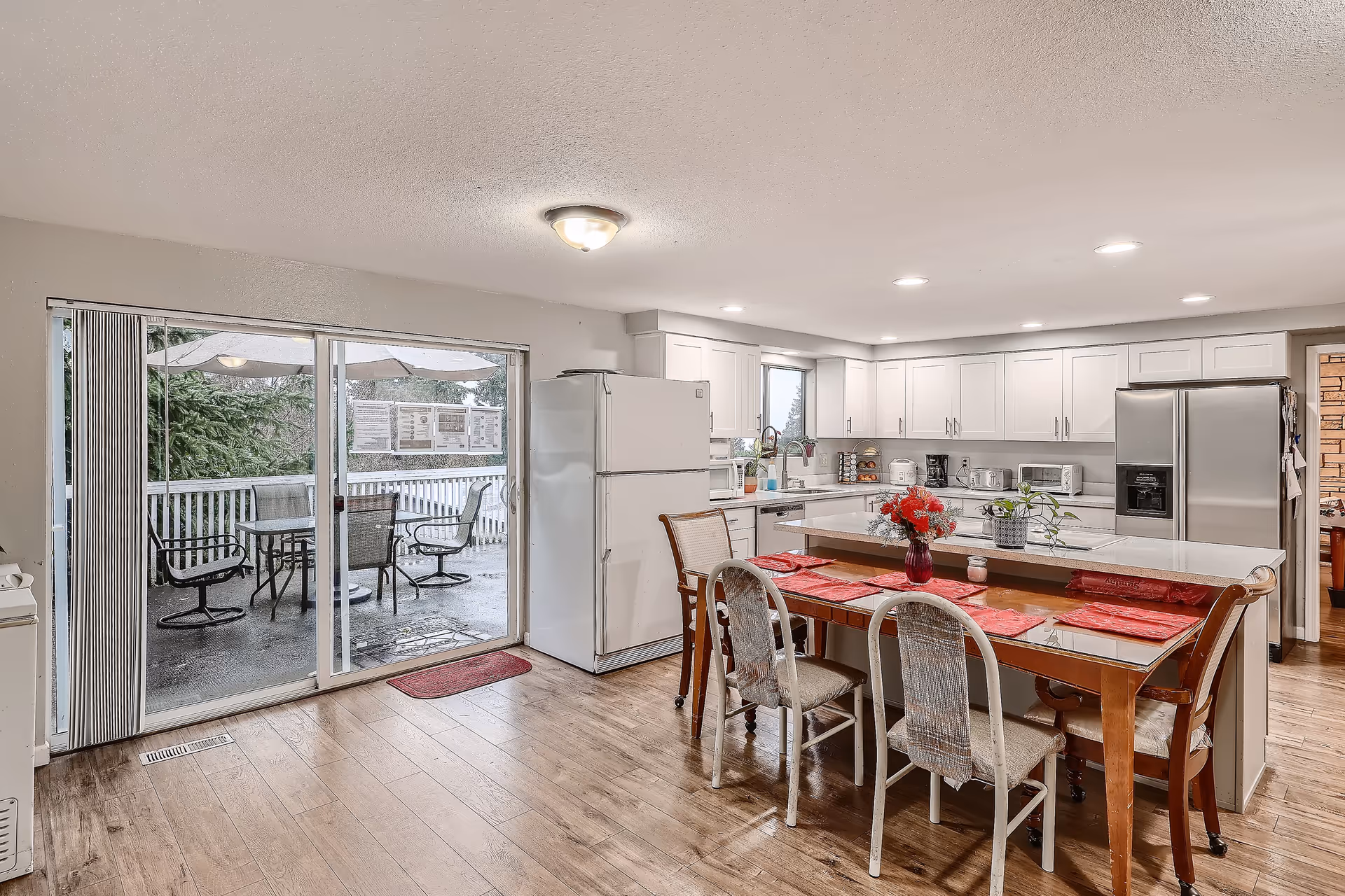 A bright kitchen and dining area with white cabinets, a white refrigerator, and stainless steel appliances. There is a wooden dining table with six chairs, each with a red placemat, and a vase with red flowers on the table. Sliding glass doors lead to an outdoor patio with a table, chairs, and an umbrella. The floor is wooden, and the ceiling has recessed lighting.