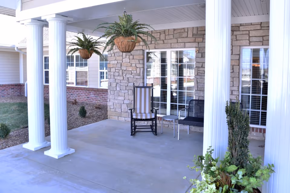 Covered outdoor patio area with white columns, two hanging fern plants, a striped rocking chair, a black chair, a small round table, and a stone wall with windows in the background.