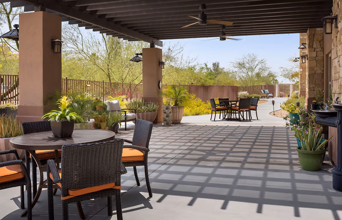 Outdoor covered patio area with round tables and wicker chairs with orange cushions, potted plants, ceiling fans, and a view of a garden with trees and shrubs under a clear blue sky.