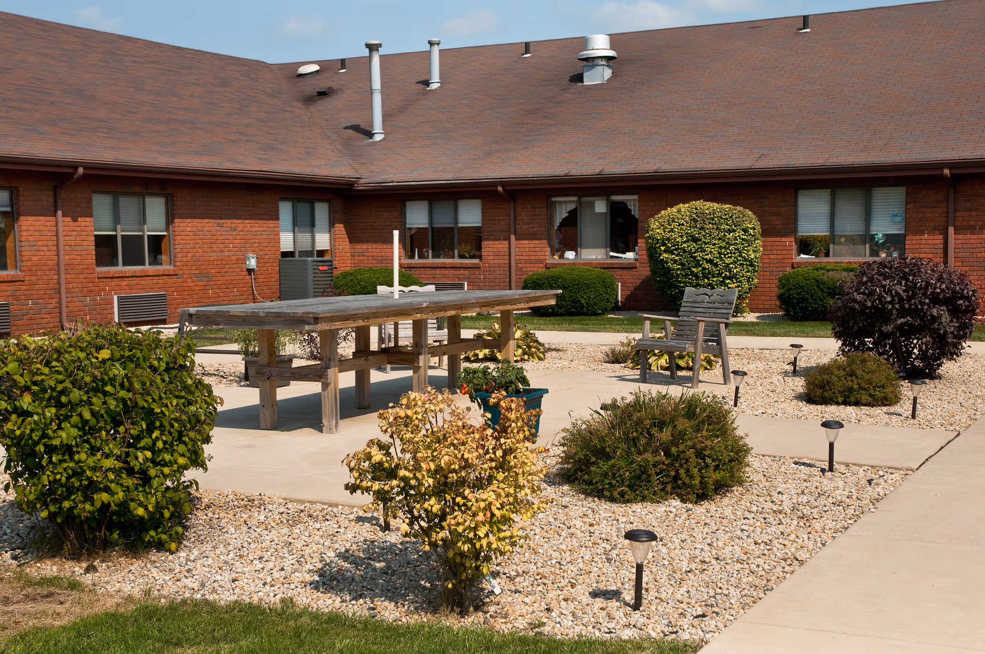 Outdoor courtyard area at Life Care Center of Valparaiso featuring a wooden picnic table, a wooden chair, various bushes and shrubs planted in gravel beds, and a brick building with multiple windows in the background under a clear sky.