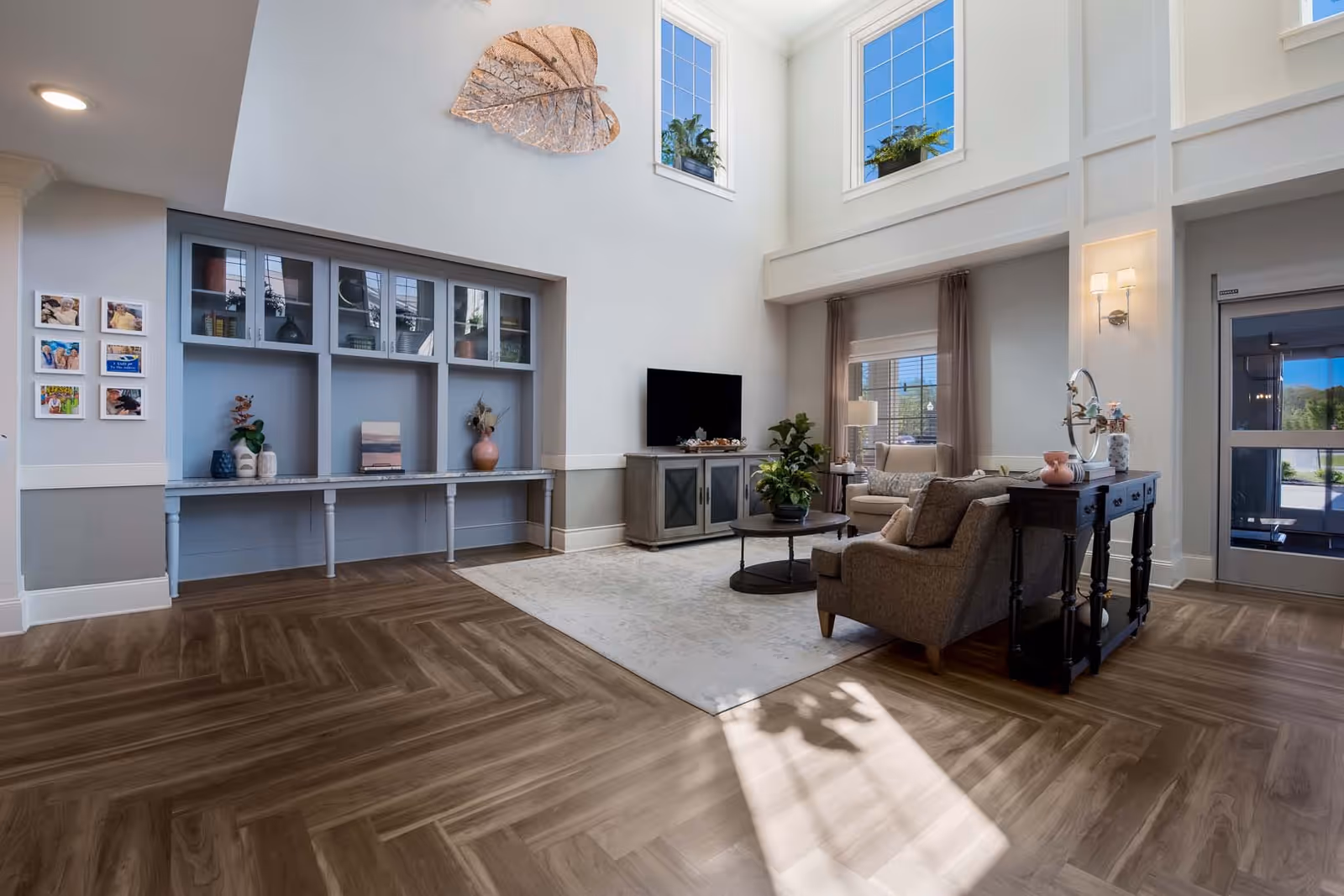 A bright and spacious living room area with high ceilings and large windows letting in natural light. The room features a comfortable seating arrangement with a sofa and armchair around a coffee table, a TV on a cabinet, and decorative plants. There is a built-in shelving unit with decorative items and framed photos on the wall. The floor has a wood pattern, and a large leaf-shaped wall decoration is mounted high on the wall.