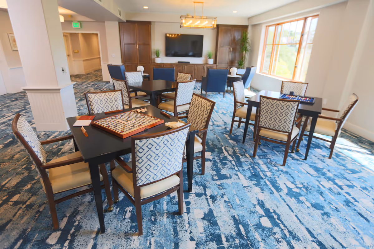 A bright and spacious common area with multiple tables and chairs arranged for games and social activities. One table has a Scrabble board game set up, and another has a backgammon board. The room features large windows letting in natural light, blue patterned carpet, a large flat-screen TV mounted on a wooden cabinet, and some potted plants.