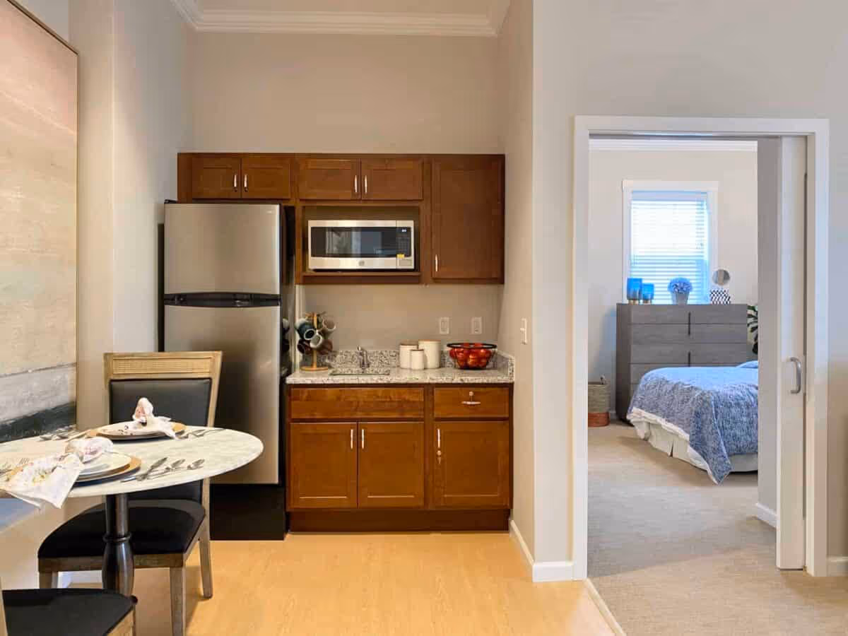 A small kitchen area with wooden cabinets, a stainless steel refrigerator, and a microwave above a granite countertop. To the left, there is a round dining table set with plates, napkins, and cutlery, accompanied by two chairs. To the right, an open doorway leads to a bedroom with a bed covered in a blue patterned bedspread and a dresser beneath a window with blinds.