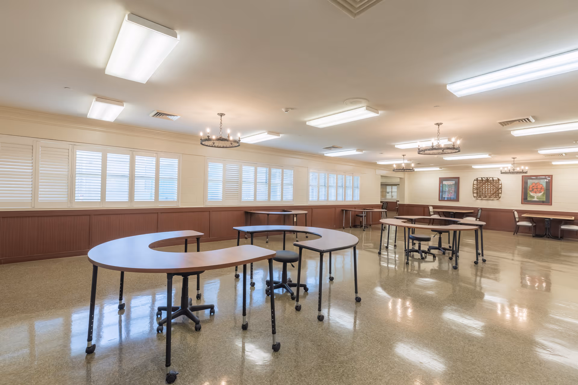 Large bright communal dining/activity room with U-shaped movable tables, stools, and shuttered windows.