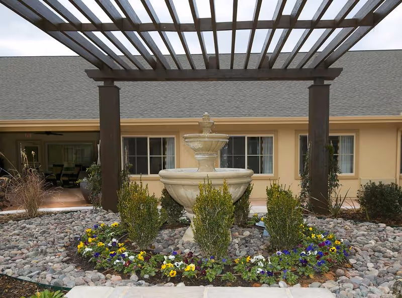 Outdoor courtyard area at Meadowbrook Memory Care Community featuring a stone fountain surrounded by small bushes and colorful flowers, with a wooden pergola overhead and the building facade in the background.