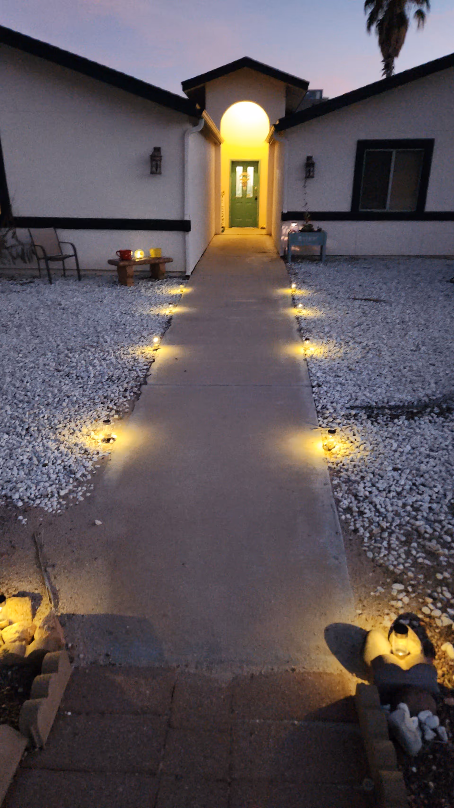 Lit walkway through gravel landscaping leading to a green front door of a single-story house at dusk.