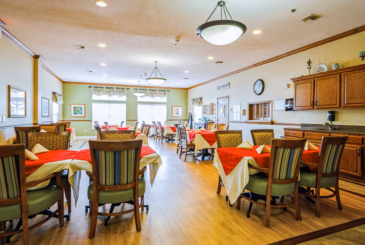 Bright dining room with multiple tables set with red and white tablecloths and wooden chairs, windows and cabinetry in the background.