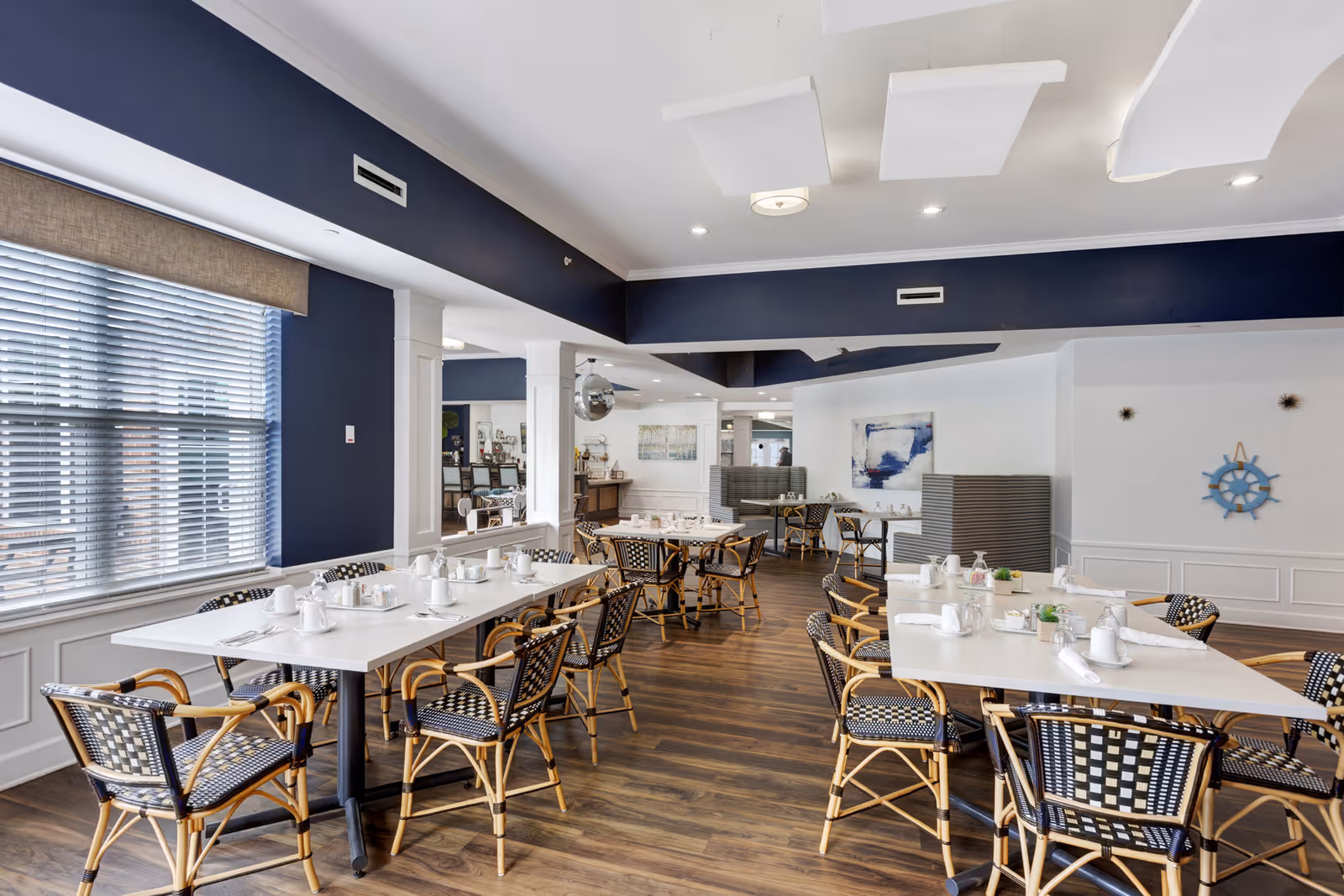 A bright and spacious dining room with multiple white tables set with white cups, plates, and napkins. The room features wooden flooring, wicker chairs with black and white cushions, large windows with blinds, and navy blue and white walls. Decorative elements include a blue ship wheel on the wall and abstract artwork.