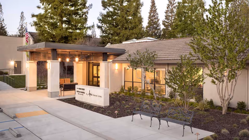 Exterior view of Atria El Camino Gardens senior living facility entrance with a covered walkway, benches, trees, and landscaping under a clear sky.