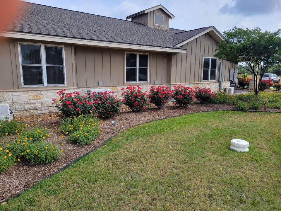 Exterior view of a single-story building with windows, a landscaped flower bed of roses and other flowers, and a grassy lawn.