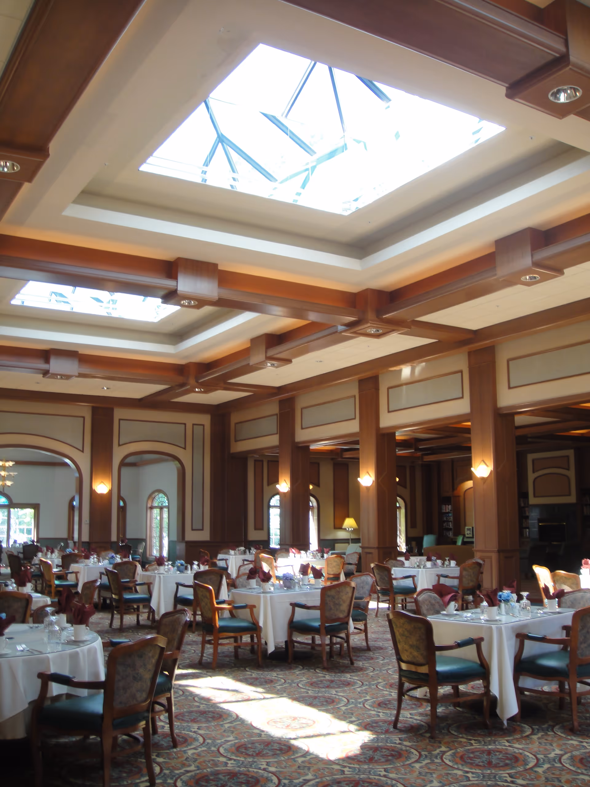 A spacious dining room with round tables covered in white tablecloths, set with glassware and napkins. The room features wooden beams on the ceiling with large skylights allowing natural light to fill the space. Upholstered chairs surround each table, and the room has decorative wall panels and arched windows.