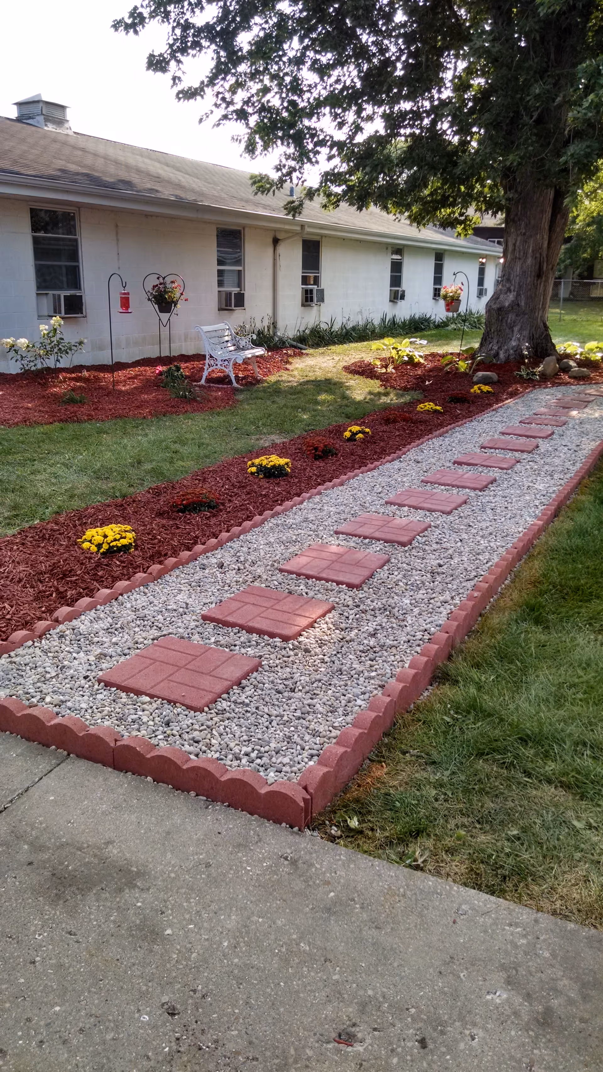 A landscaped garden area outside a single-story building with white walls and several windows. The garden features a gravel pathway with red square stepping stones bordered by red bricks. There are flower beds with red mulch and yellow flowers, a white bench, hanging flower baskets, and a large tree providing shade.