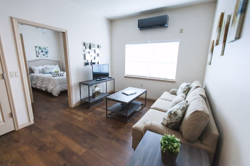 A bright living room with a beige sofa adorned with patterned pillows, a coffee table, and a TV on a stand. The room has wooden flooring and a large window with blinds. An air conditioning unit is mounted above the window. Through an open doorway, a bedroom with a neatly made bed and wall art is visible.