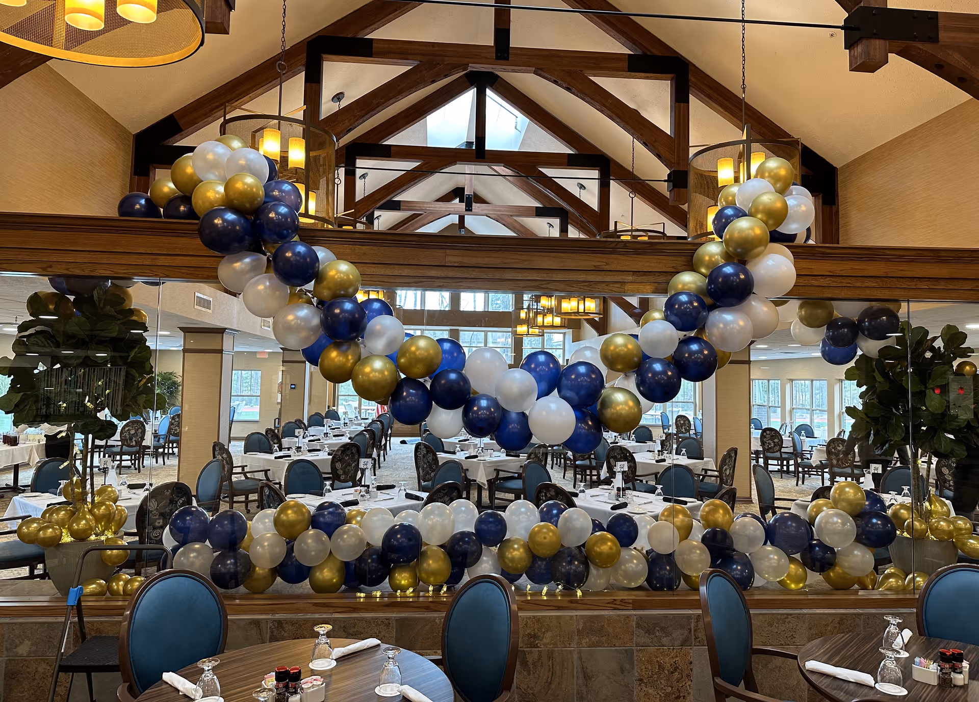 Dining room decorated with blue, gold, and white balloon garlands reflected in a large mirror, with tables and chairs set for a meal.