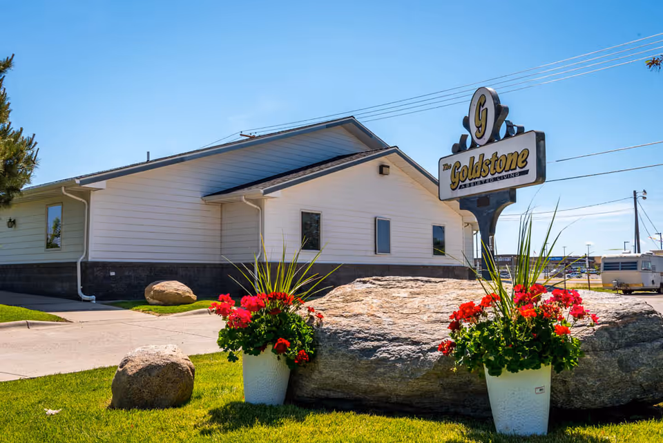 Exterior view of The Goldstone Assisted Living building with a sign displaying the facility's name. The building is white with a dark stone base, and there are large rocks and flower pots with red flowers in the foreground under a clear blue sky.