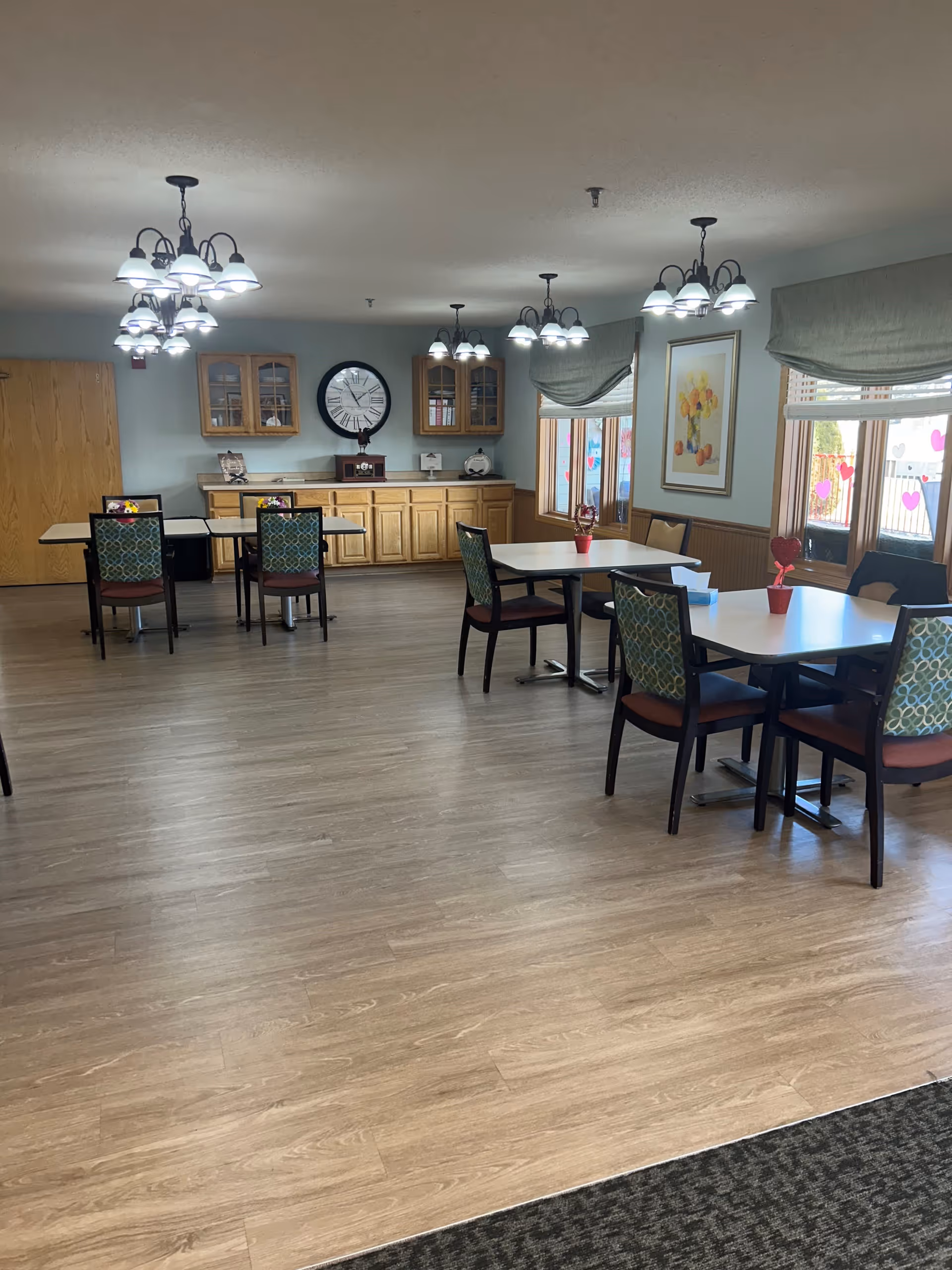 A dining room in an assisted living facility with several tables and chairs arranged neatly. The room has wooden flooring, light blue walls, and multiple ceiling light fixtures. There are windows with green valances letting in natural light, a large wall clock, wooden cabinets, and framed artwork on the walls. Small decorative items are placed on the tables and cabinets.