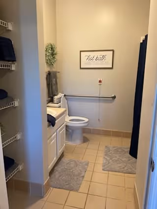 A clean and accessible bathroom with beige tiled floor, a white toilet, a white vanity with a sink, and two gray bath mats on the floor. There is a grab bar next to the toilet and a pull cord emergency alarm on the wall. Towels are neatly folded on shelves to the left, and a framed sign on the wall reads 'Hot bath'.