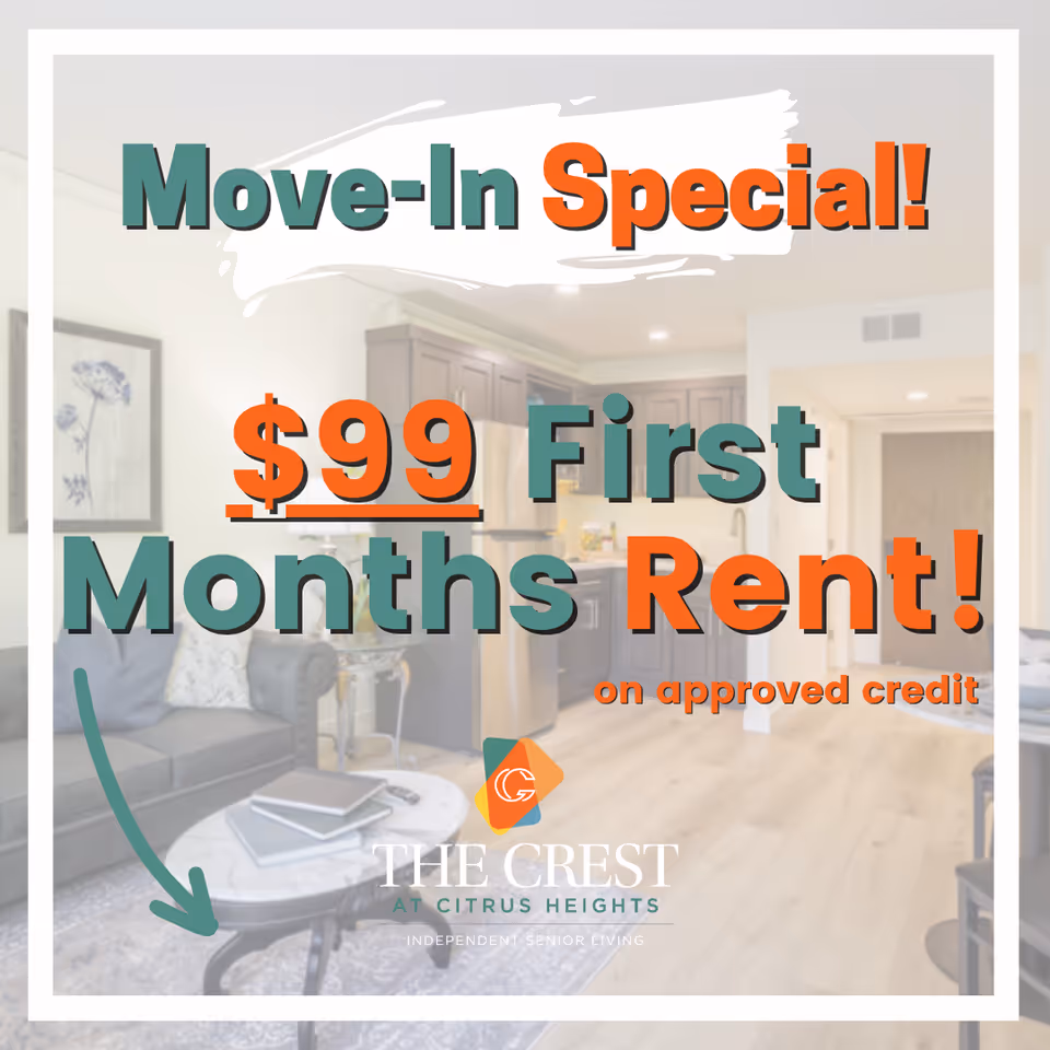 Interior view of a senior living facility showing a living room area with a gray sofa, a round coffee table with books, and a kitchen in the background with wooden cabinets and stainless steel appliances.