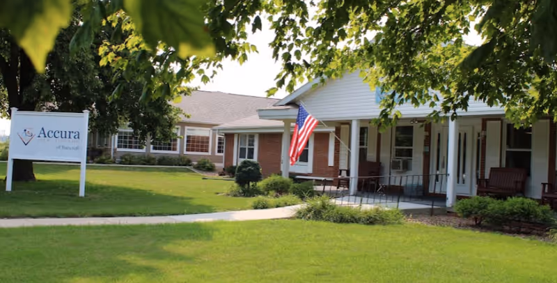 Exterior view of Accura Healthcare of Bancroft building with a well-maintained lawn, a sidewalk, an American flag hanging near the entrance, and a sign displaying the facility's name.