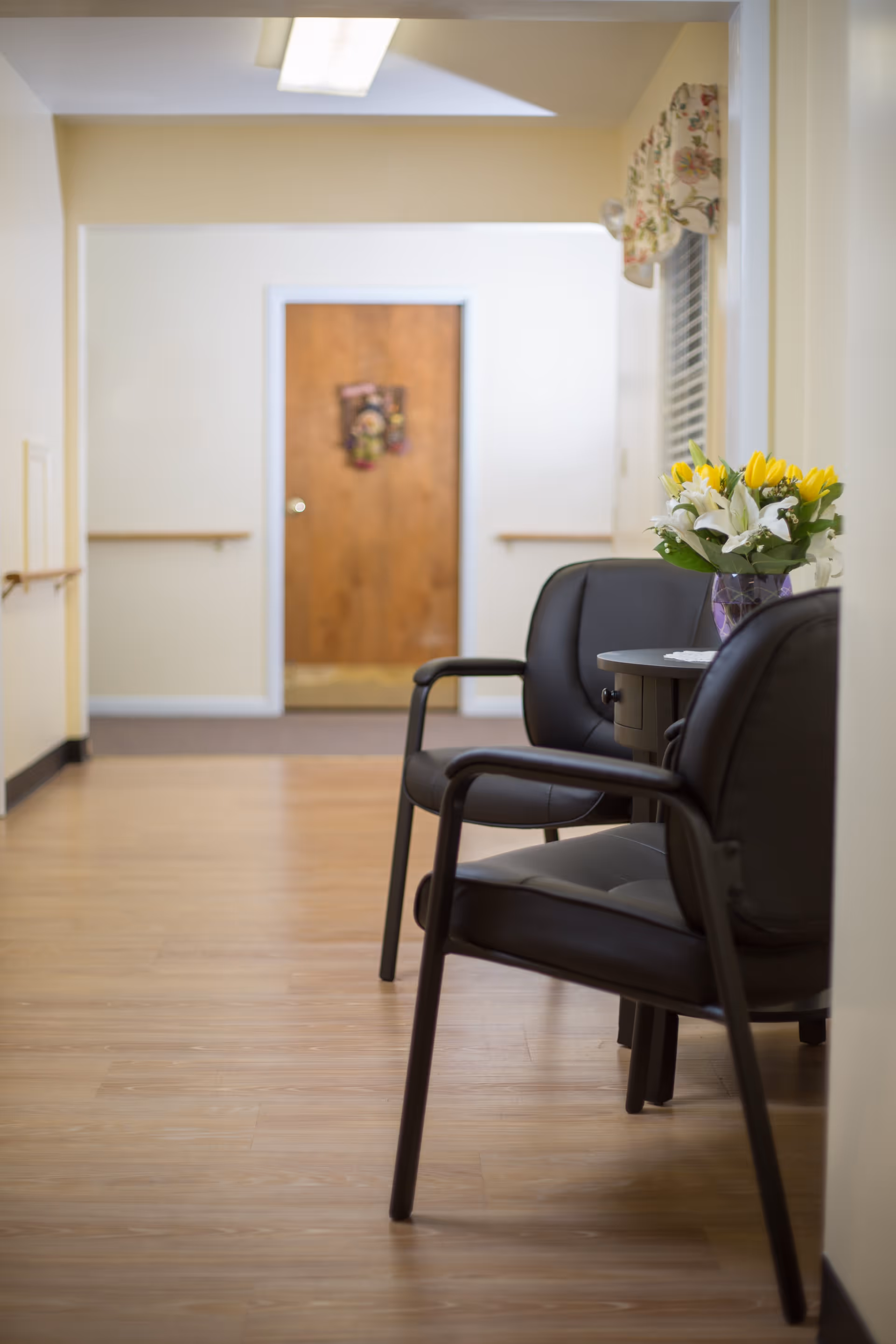 A hallway with light-colored walls and wooden flooring featuring two black chairs and a small round table with a vase of yellow and white flowers. A wooden door with a decorative wreath is visible at the end of the hallway.