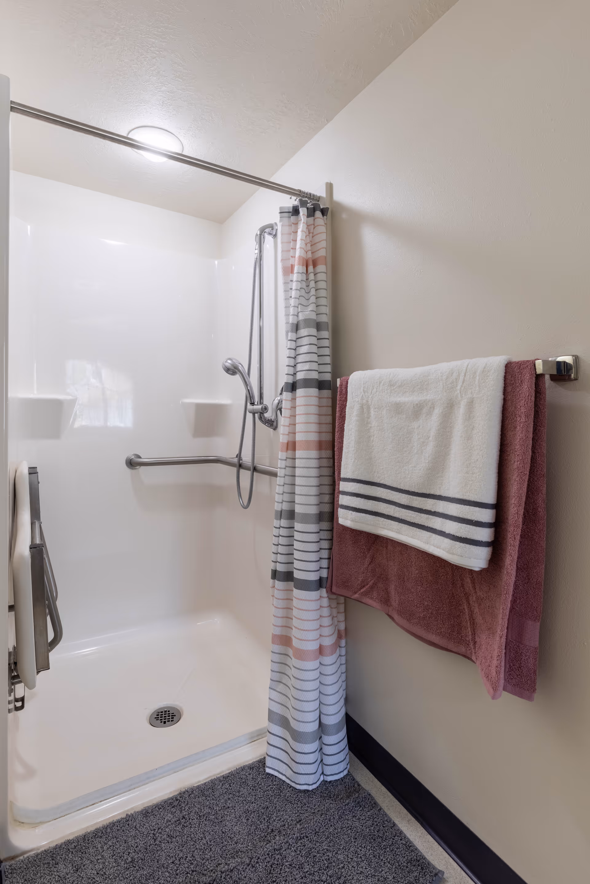 A clean, accessible shower area with a white shower curtain featuring horizontal stripes in gray, white, and pink. The shower has a handheld showerhead, grab bars, and a fold-down seat. Two towels, one white with gray stripes and one dark pink, hang on a towel rack mounted on the beige wall next to the shower. A gray bath mat is placed on the floor outside the shower.
