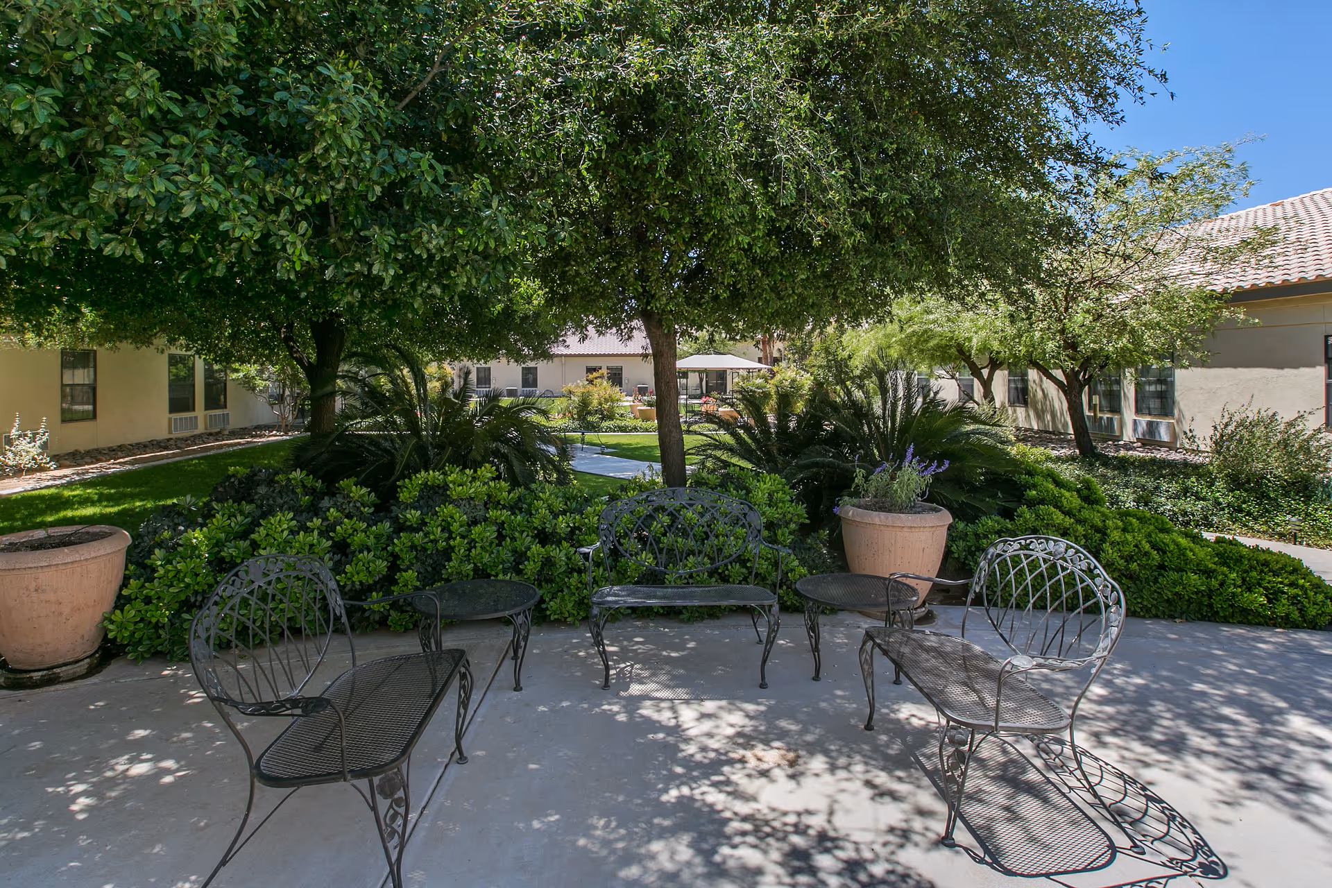 Shaded outdoor courtyard with metal patio chairs and tables surrounded by trees, shrubs, potted plants, and adjacent single-story buildings.