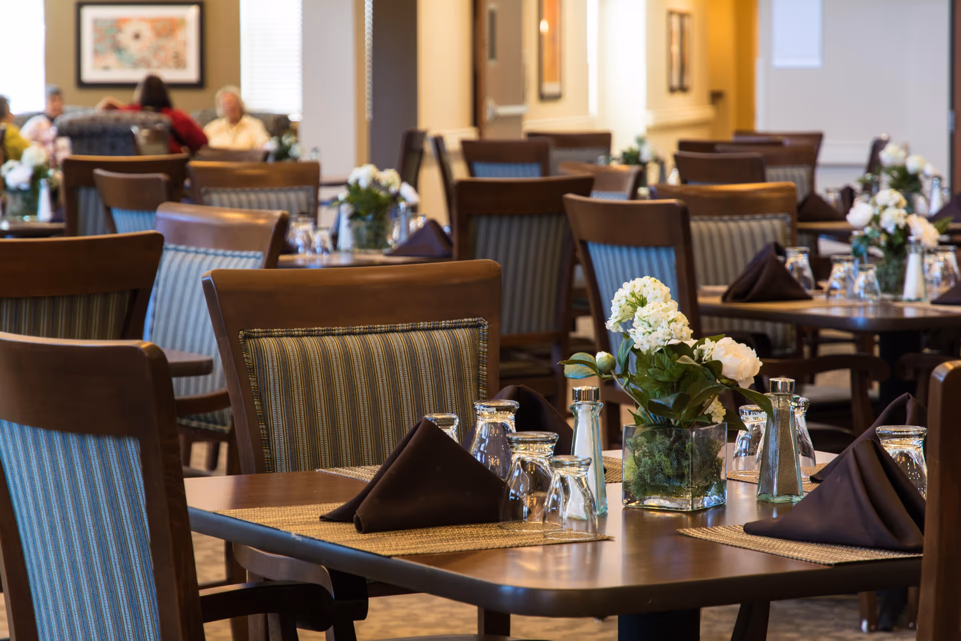 A dining room with multiple wooden tables and chairs, each table set with brown napkins, glassware, salt and pepper shakers, and floral centerpieces. In the background, a few people are seated and engaged in conversation.